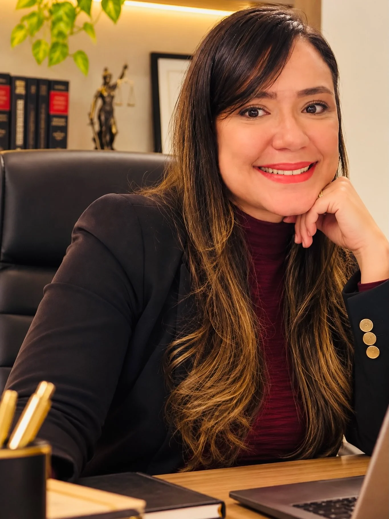 Dra. Tielle Menezes, advogada, sorrindo com a mão apoiada no queixo, sentada à mesa do escritório.